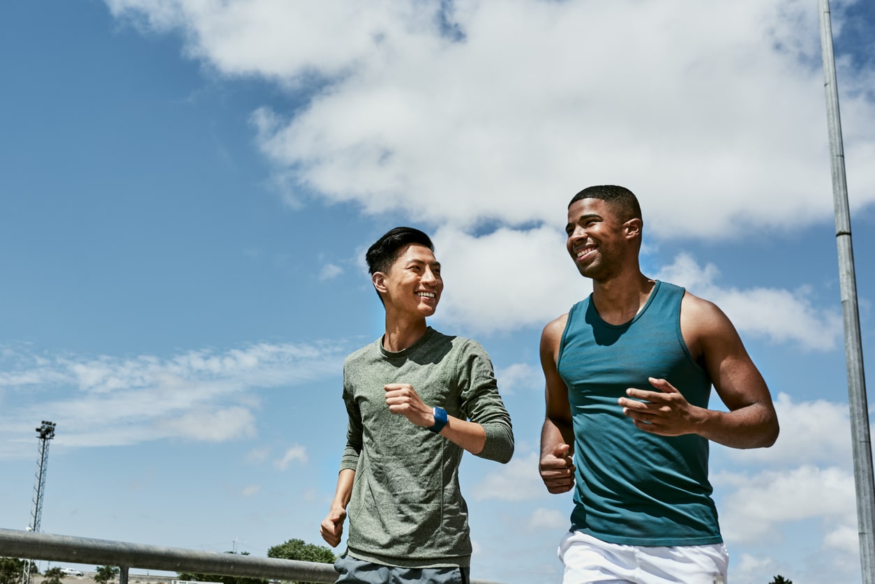 Active men jogging outdoors on blue cloudy sky. Two athletic guys or young sports friends running together, doing their routine cardio workout and fitness exercise in the city